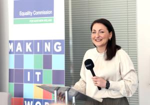 Photograph of the Ema Cubitt the Autism Reviewer at a metal podium with a microphone in her hand.  She is standing beside a stand with the Equality Commission for Northern Ireland logo on it.